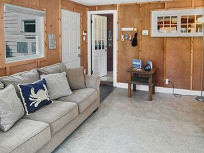 A cozy living room with a beige sofa, decorative pillows, a small wooden side table, and a wall-mounted rack with beach-themed items.