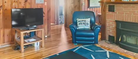 Een gezellige kamer met een televisie op een houten standaard, een blauwe leren fauteuil met een kussen, een open haard en een blauw vloerkleed.