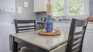 Une table à manger agrémentée d’un set de table en fibres tressées et d’un vase contenant des plantes séchées, le tout placé dans une cuisine aux meubles blancs et dotée d’une fenêtre avec des stores.