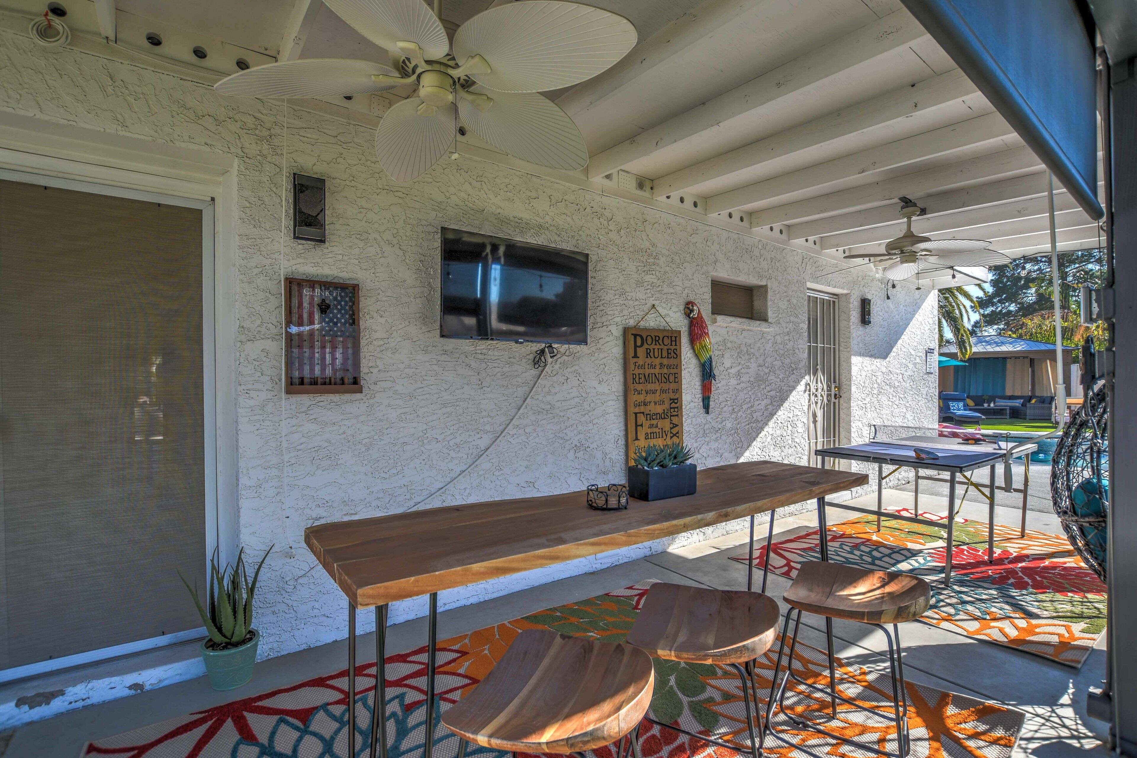 A covered patio area with a wooden table and chairs, a ceiling fan, and a TV mounted on the wall.