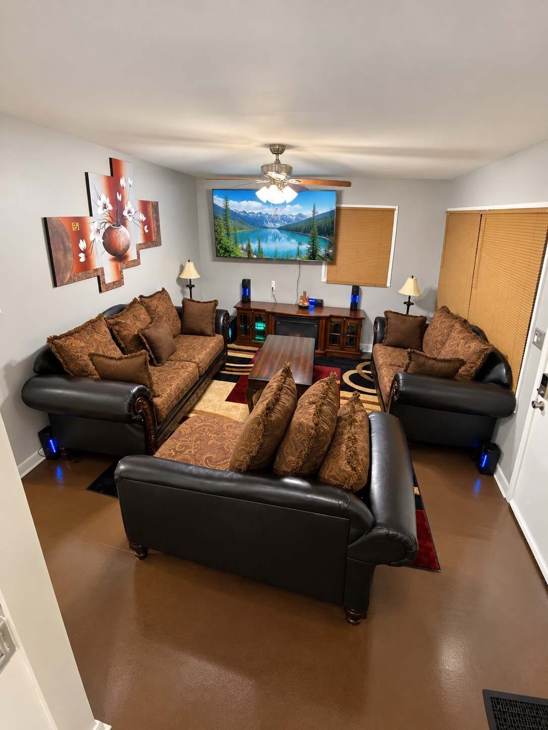 A living room with a black leather sofa set, a flat-screen TV mounted on the wall, a ceiling fan, and a fireplace.