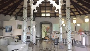 Ampla sala de estar com teto alto, colunas brancas e um lustre central. Há uma mesa de jantar com cadeiras, um sofá e uma cozinha ao fundo.