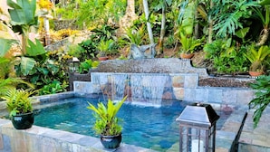 Uma piscina retangular com cachoeira, rodeada por paredes de pedra e vegetação exuberante.