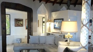 Uma sala de estar ampla com um sofá seccional, uma mesa de centro de vidro e uma poltrona branca. Há um abajur sobre uma mesinha de apoio e um quadro na parede.