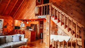 Una cabaña acogedora con interior de madera, una escalera, una cocina y una sala de estar con sofá y alfombra blanca y mullida.