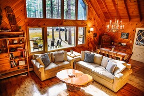 Relax in a cozy cabin-style space with a central seating area, wooden bookshelf, and large windows framing the surrounding forest.