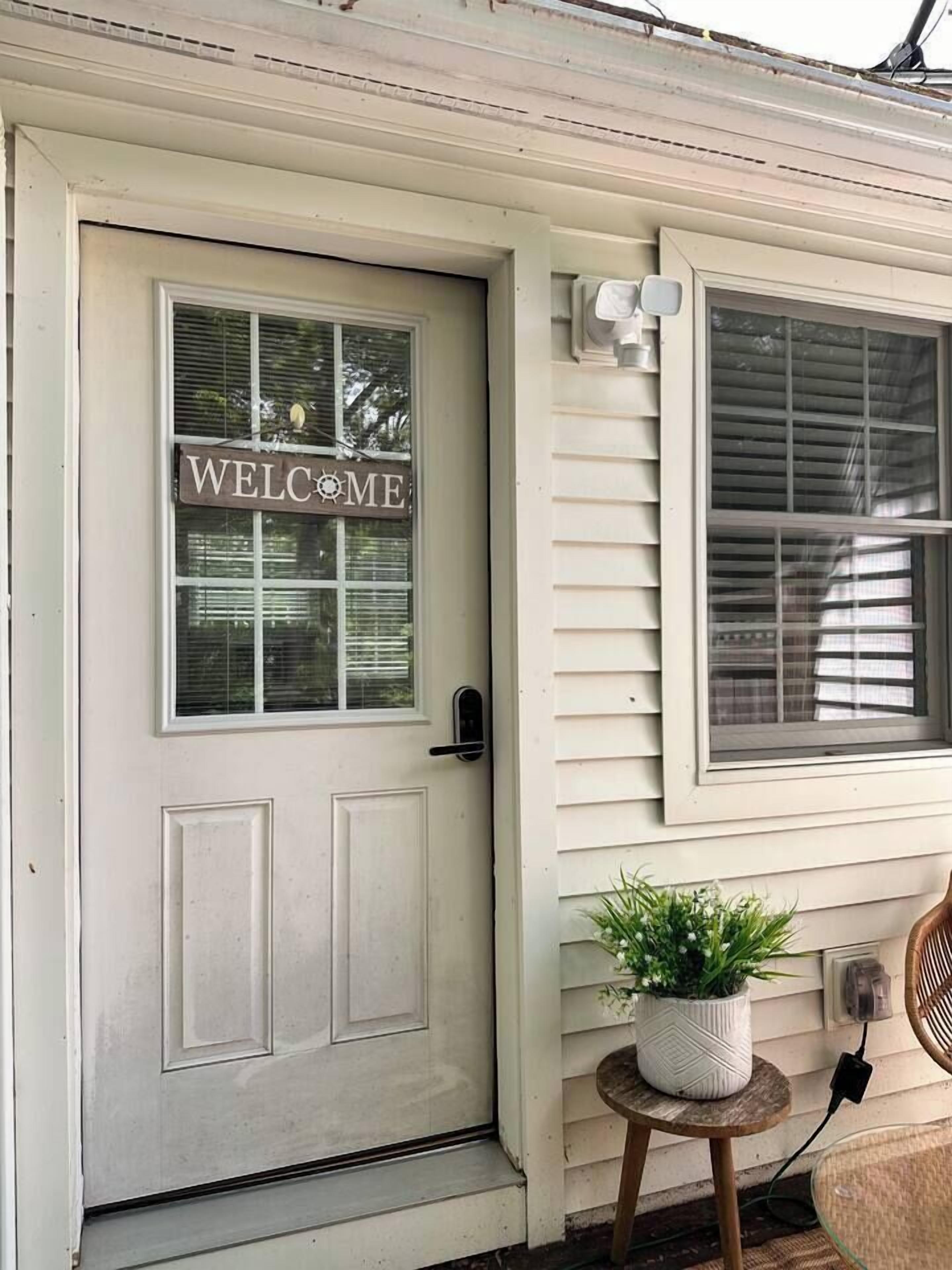 A white door with a 'WELCOME' sign, a small potted plant on a wooden stool, and a window with blinds.