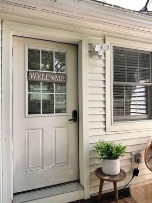 A white door with a 'WELCOME' sign, a small potted plant on a wooden stool, and a window with blinds.