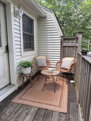 A wooden deck with two wicker chairs and a small table, a potted plant, and a door to a house.