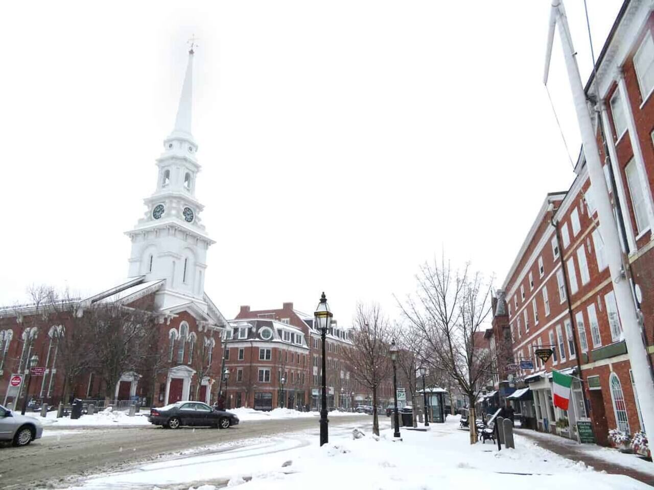 A snow-covered street with historic buildings, a prominent white church steeple, and a flag.