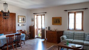 Ein gemütliches Wohnzimmer mit einem Holzesstisch, Stühlen, einer Couch und einem Couchtisch. Es gibt eine Chandelier, ein Bild an der Wand und ein Fenster mit Vorhängen.