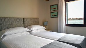 Een tweepersoonsbed met witte lakens, een hoofdbord met patroon, een nachtkastje met een lamp, en een raam met uitzicht op zee.