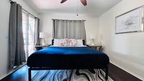 Ein Schlafzimmer mit einem Bett, zwei Nachttischen mit Lampen, einem Deckenventilator, einem Fenster mit Vorhängen und einem gerahmten Bild an der Wand.