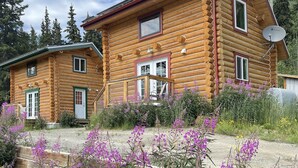 A two-story log cabin with a satellite dish, a front porch, and a garden with purple flowers. - Log Cabin with Amazing Mountain & Lake Views  (allergy-friendly) (Carcross)