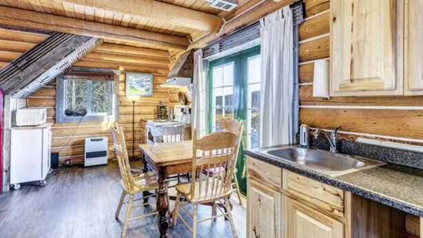 A wooden cabin interior with a dining area, kitchen, and a large window. - Log Cabin with Amazing Mountain & Lake Views  (allergy-friendly) (Carcross)