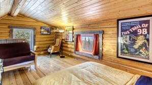 A cabin interior with wooden walls, a bed, a sofa, a chair, and a poster of 'The Trail of 98'. - Log Cabin with Amazing Mountain & Lake Views  (allergy-friendly) (Carcross)