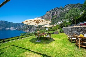 Un patio con mesa y sillas, un gran paraguas y vistas al mar y a las montañas.