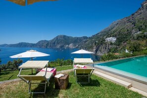 Zona junto a la piscina con dos tumbonas y sombrillas, con vistas a un mar azul cristalino y un paisaje montañoso.