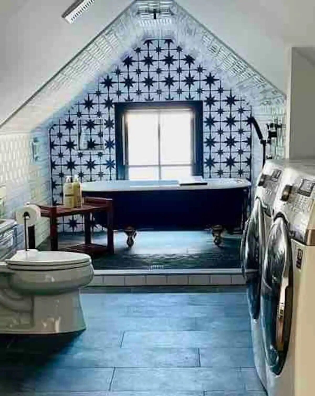 A small, sloped-roofed bathroom with a star-patterned tile wall, a window, a bathtub, a toilet, and a washing machine.