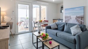 Ett modernt vardagsrum med en grå soffa, ett träbord som används som soffbord och som är dekorerat med blommor, en matplats med bord och stolar samt ett köksområde med en spis och skåp.