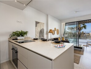 A modern kitchen with a large island, a dining area with a glass table, and a view of the outdoors through large windows.