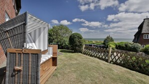 A cozy outdoor seating area with a wicker chair and a cushioned bench, overlooking a grassy lawn and a view of the sea. - Lena's Hüs - Beautiful thatched house for 6 pers. with sea view, garden, WLAN, (List)