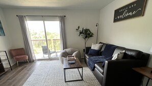 Ein modernes Wohnzimmer mit einer Couch, einem Sessel und einem Couchtisch, eine Schiebetür mit Glas zum Balkon und ein Schild mit der Aufschrift 'Hello and Welcome'.