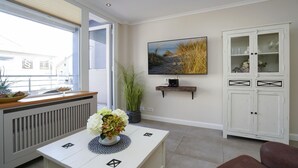Ein modernes Wohnzimmer mit einer weißen Heizung, einem weißen Couchtisch mit geometrischem Design, einem großen Flachbildfernseher an der Wand, einer weißen Vitrine mit Glastüren und einer Topfpflanze.