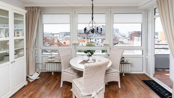 Ein Essbereich mit einem runden Tisch, Korbstühlen und Blick auf die Stadt durch große Fenster.