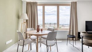 Una sala de estar moderna con una mesa redonda de madera, sillas tapizadas en gris, una lámpara de pie, un televisor y vistas de edificios a través de un gran ventanal con cortinas beige.