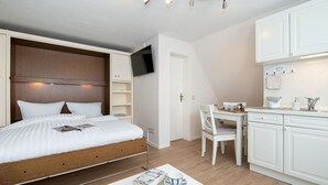 Una habitación compacta con cama, mesitas de noche, televisor y una pequeña kitchenette con zona de comedor.
