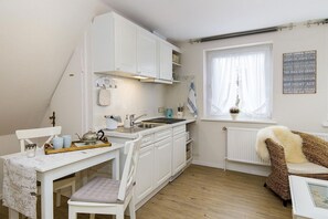 Et kompakt køkken med hvide skabe, et lille spiseområde med et bord og stole, et vindue med gardiner og en fletstol.