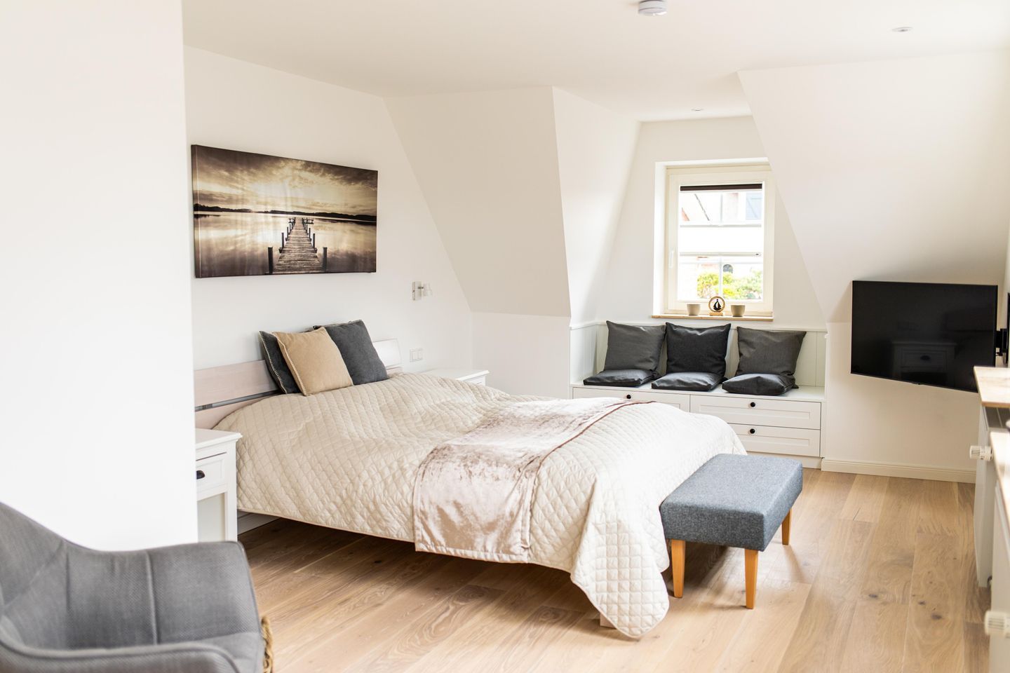Un dormitorio moderno con cama, mesitas de noche, un banco, televisor y ventana con vistas.