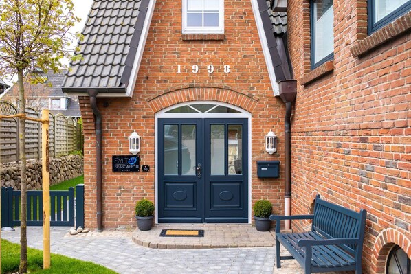 Un bâtiment en briques avec une porte d’entrée bleu foncé, une enseigne indiquant « SYLT SEASCAPE » et un banc à l’extérieur.