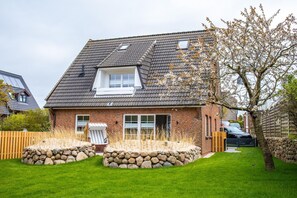 Une maison à deux étages, dotée d’un toit en pente, d’un porche à l’avant et d’un jardin bordé d’un mur en pierre.