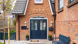 A brick building with a dark blue front door, a sign reading "SYLT SEASCAPE," and a bench outside.