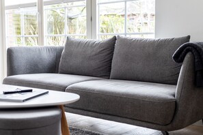 Un canapé gris avec une télécommande posée dessus, une petite table ronde sur laquelle se trouvent un livre et un stylo, et une fenêtre donnant sur un paysage verdoyant.