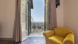 A room with a yellow armchair, an open door leading to a balcony with a view, and a wall-mounted clock.