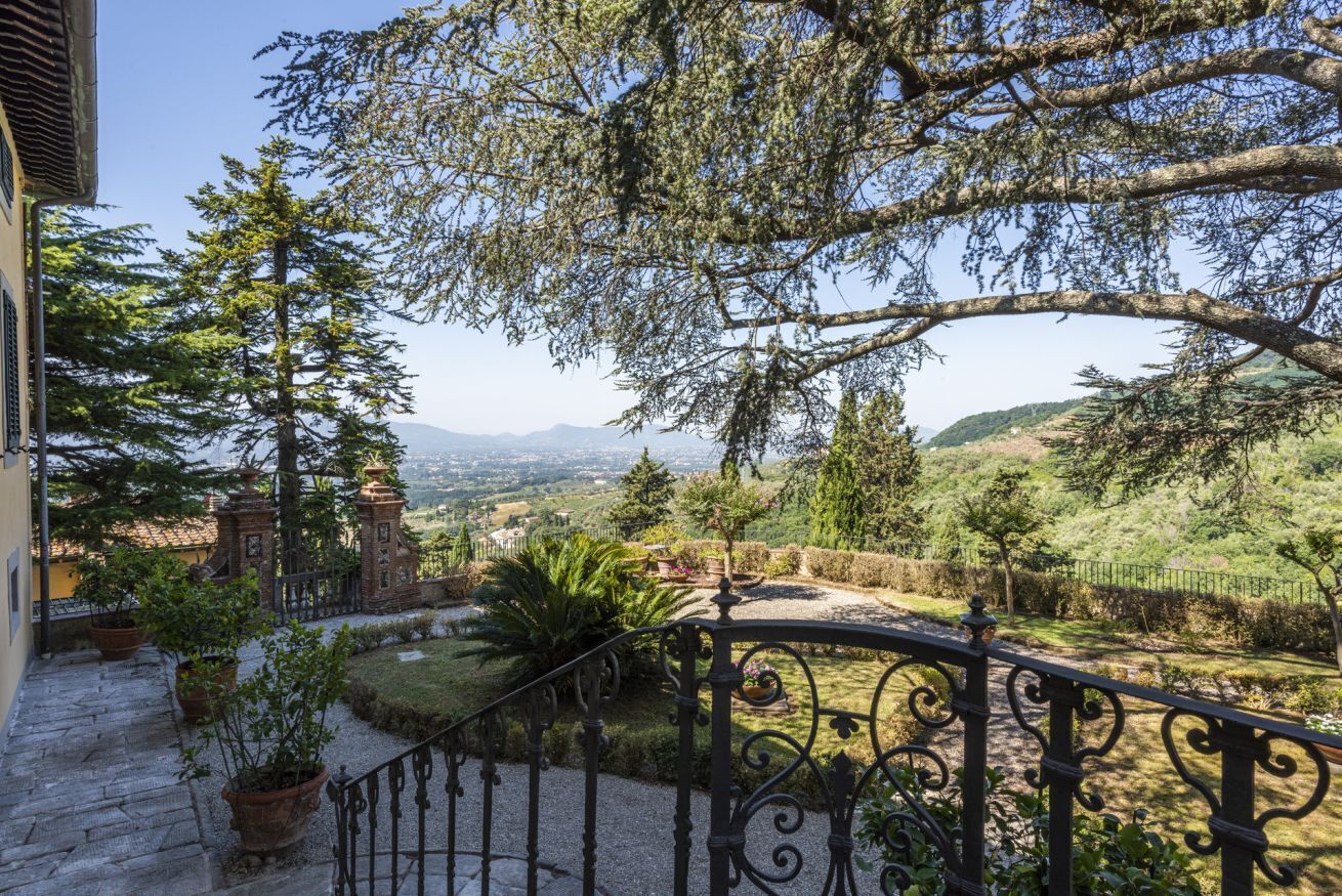 A stone staircase leading to a balcony with a wrought iron railing overlooking a landscaped garden and distant hills.