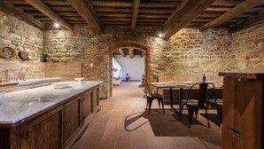 A rustic kitchen with stone walls, wooden beams, a marble countertop, and a dining area with chairs.