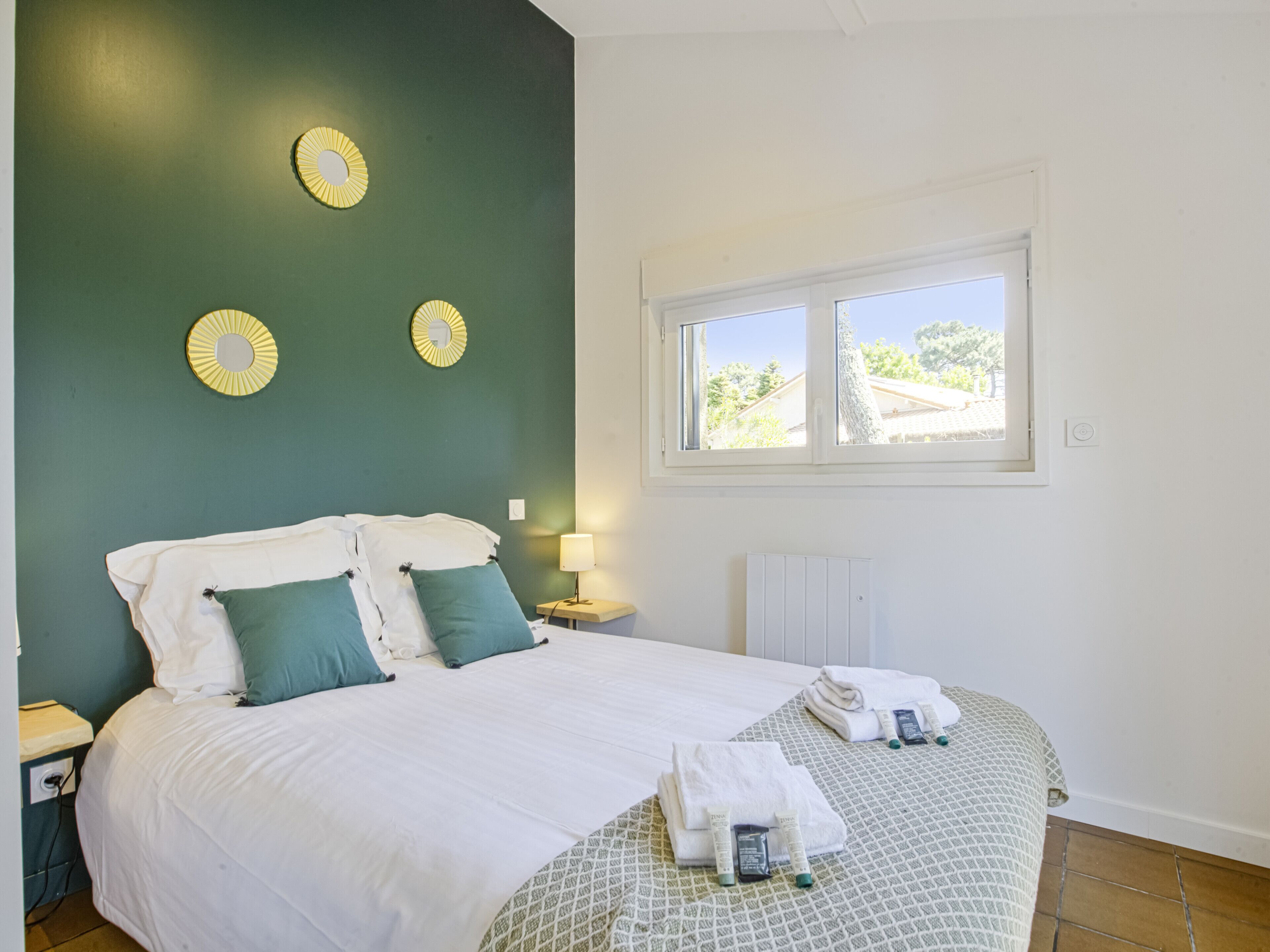 Un lit bien fait, avec une literie blanche et verte, une fenêtre donnant sur des arbres et un mur orné de miroirs circulaires jaunes.