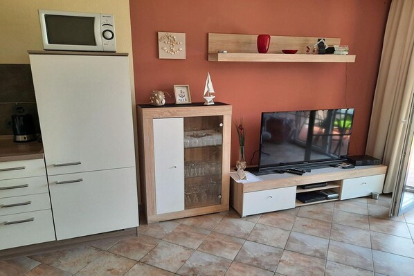Een ruimtelijk ingerichte leefruimte met een kitchenette, een televisie en een houten kast met glazen deuren.