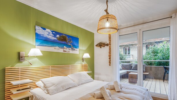 A bedroom with a bed, bedside tables, a painting of a beach scene, a hanging light fixture, and a view of a balcony with outdoor furniture. - Appartement Thiers - Welkeys (Antibes)