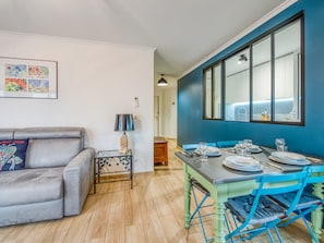 Una sala de estar moderna con un sofá gris, una mesita con una lámpara, y un comedor con una mesa y sillas azules.