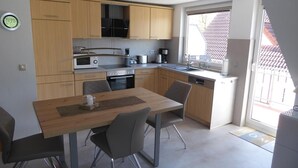 A modern kitchen with wooden cabinets, a microwave, an oven, and a sink. There is a dining table with chairs and a window providing a view of the outside. - Vacation apartment Am Harlebogen 11 Whg. 4 (Wittmund)