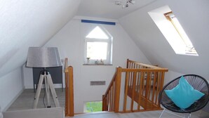 A spacious attic room with a sloped ceiling, a wooden staircase, a tripod lamp, a comfortable chair, and a skylight. - Vacation apartment Am Harlebogen 11 Whg. 4 (Wittmund)