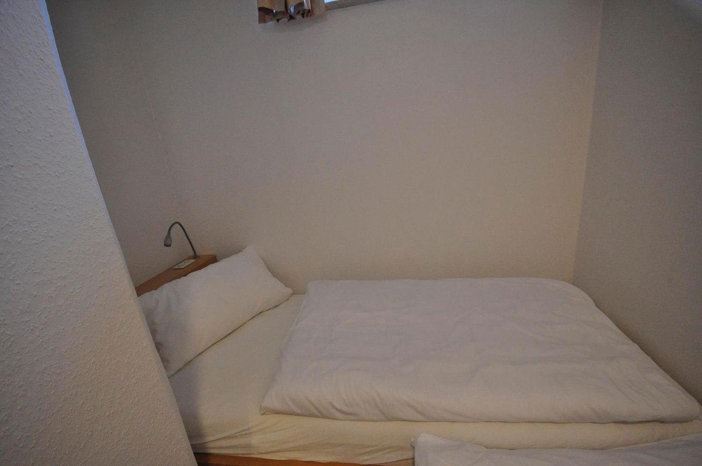 Une petite chambre avec un lit simple, dotée de draps blancs et d’une tête de lit en bois.