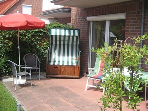 A patio area with a striped beach chair, a red umbrella, and a small table with a green tablecloth. - Vacation apartment Am Harlebogen 10 Whg. 2 (Wittmund)