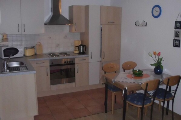 A compact kitchen with a stove, oven, and sink, a dining table with chairs, and a clock on the wall. - Vacation apartment Am Harlebogen 10 Whg. 2 (Wittmund)