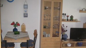 Zona de menjador amb taula, cadires, un armari amb vetrereria, televisió i una petita estant amb objectes decoratius.